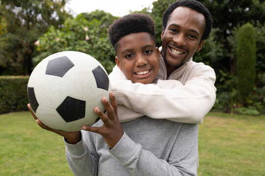 African American dad hugging teen son on lawn holding black-and-white soccer ball, gray sweatshirt