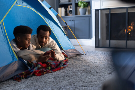African American father and teen son sharing smartphone in blue-yellow pop-up tent in living room