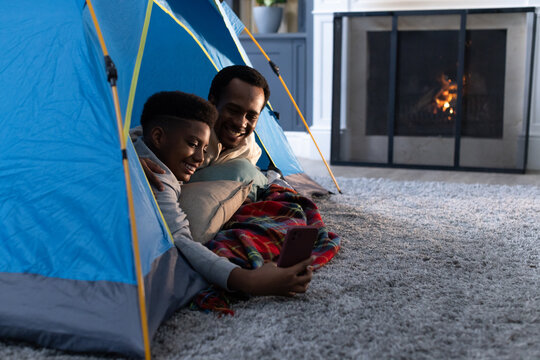 African American father and teenage son laughing, looking at smartphone inside blue tent on carpet