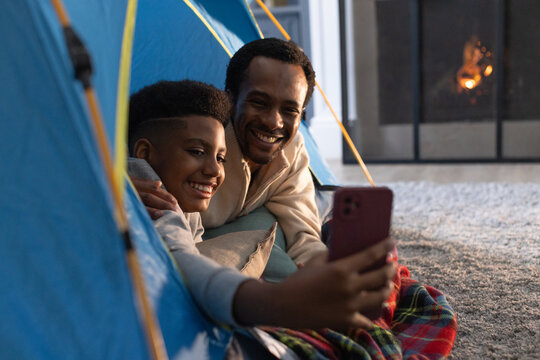 African American father and adolescent son taking selfie with phone inside blue tent by fireplace
