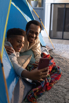 African American father and teen son smiling while watching smartphone in blue popup tent on carpet
