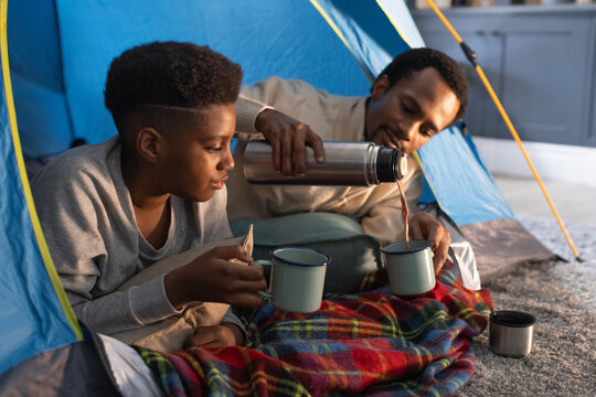 African father, teen son reclining in blue tent at home, pouring from steel thermos into mugs