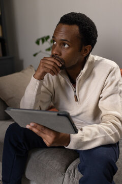 African American male sitting on gray sofa at home holding tablet in folio case wearing pullover