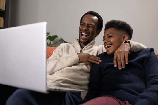 African American father and teenage son sitting on sofa laughing at laptop with throw pillow