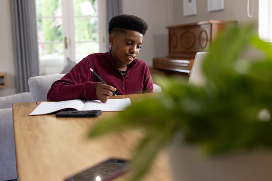 African American teen sitting at wooden table at home writing in open notebook with pencil, piano