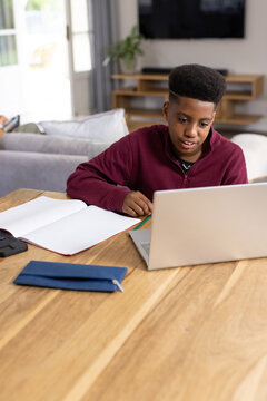 African American teenage male studying at dining table at home using silver laptop and notebook