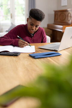 African American teen doing homework at home table in maroon zip-up with notebook and green pencil