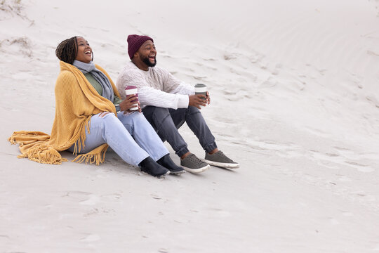 African American couple sitting on dunes holding coffee cups wearing mustard blanket, copy space