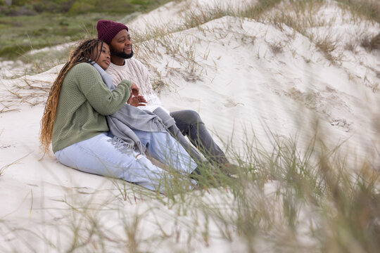 African American couple sitting on sand dune, leaning, wearing knitwear beanie and gray blanket