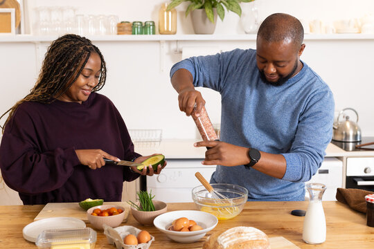 African couple slicing avocado and seasoning beaten eggs at wooden countertop, wearing sweaters