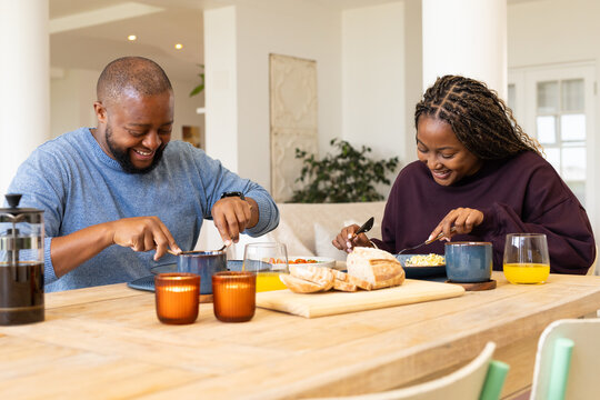 African American couple in blue and maroon sitting at dining table cutting bread with French press