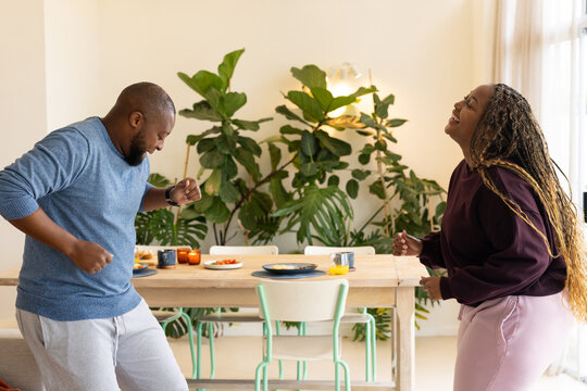 African American couple dancing and laughing at home beside wooden dining table with orange juice