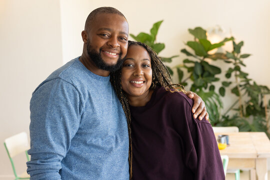 African couple smiling in dining room wearing blue sweater and plum sweatshirt with wooden table