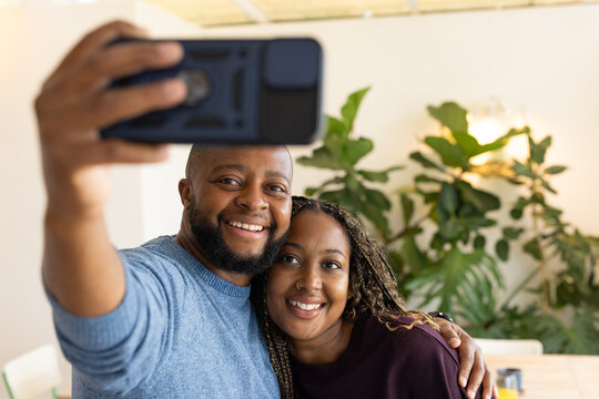 African couple posing and taking selfie at home wearing blue sweater, maroon top with smartphone