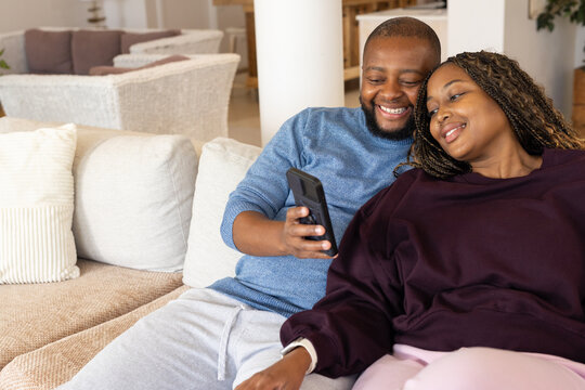 Couple sitting closely together on light-colored fabric sofa in living room, looking at smartphone
