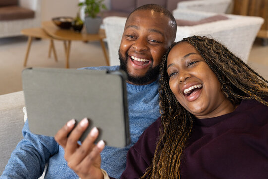 African American couple laughing and sharing gray tablet on sofa in blue knit and plum sweatshirt