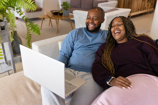 African couple sitting on light sofa laughing and watching silver laptop at home with smartwatch