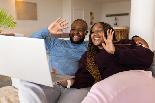 Couple wearing sweaters sitting on sofa waving at laptop on lap in living room with plant