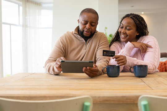 Couple sitting at dining table in knits, using tablet while woman pointing holding credit card