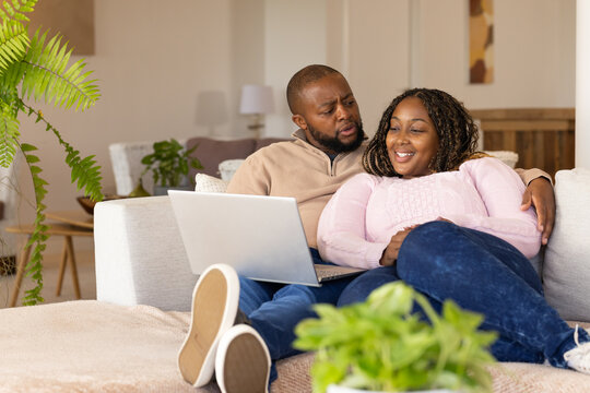 African couple sitting on light sofa, man holding silver laptop, woman wearing pink sweater smiling
