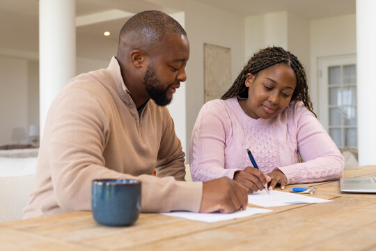 African American couple in knit sweaters sitting at dining table signing paperwork with pen, laptop