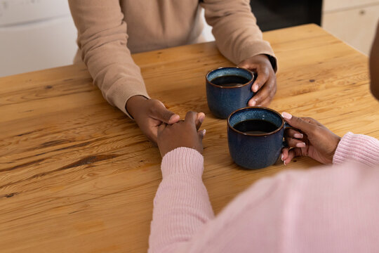 Two hands and forearms are resting and holding blue mugs on wooden dining table near cabinets