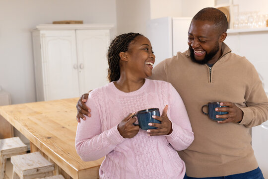 Couple laughing, holding dark-blue mugs, in pink knit and tan pullover by wood island in kitchen
