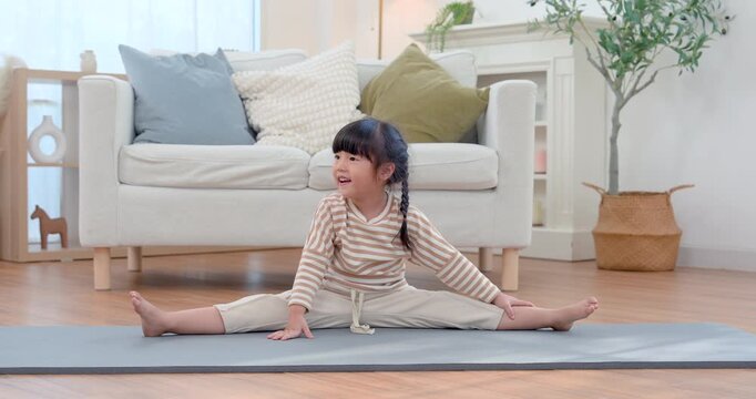 Asian preschool girl smiles during wide leg yoga stretch on exercise mat in bright living room, tilting head with playful energy while building flexibility, balance, strength, body control, wellness.