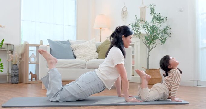 Asian single mother and preschool daughter lift through cobra pose on mat during home yoga, sharing active wellness routine that improves flexibility, body control, confidence, bonding, development