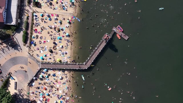 Aerial drone view of a crowded summer lake beach with people swimming and sunbathing. Colorful umbrellas, water obstacle course on lake, on a sunny day. Lake Pogoria in Dabrowa Gornicza, Poland