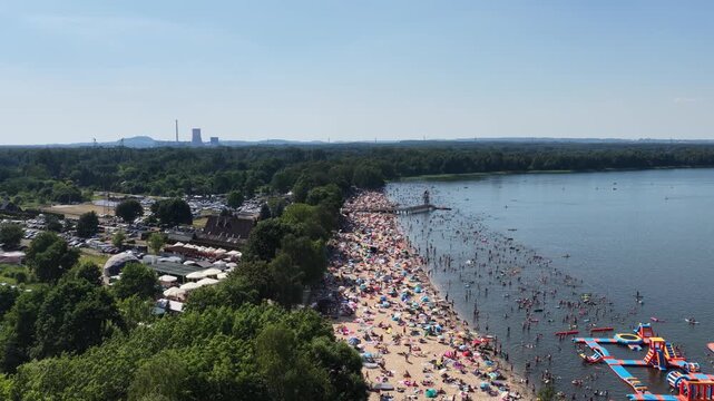 Aerial drone view of a crowded summer lake beach with people swimming and sunbathing. Colorful umbrellas, water obstacle course on lake, on a sunny day. Lake Pogoria in Dabrowa Gornicza, Poland