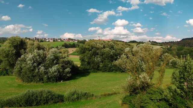 Flying towards a German village on the horizon on beautiful green hills, with trees, blue sky and fluffy white clouds in the background