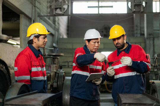 Senior engineer holding tablet explaining and inspecting machine part with technician team in factory.