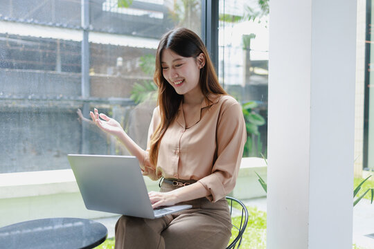 Smiling young woman using a laptop by a large window in a bright cafe or office, working online in a relaxed modern space with natural light and a cheerful mood.