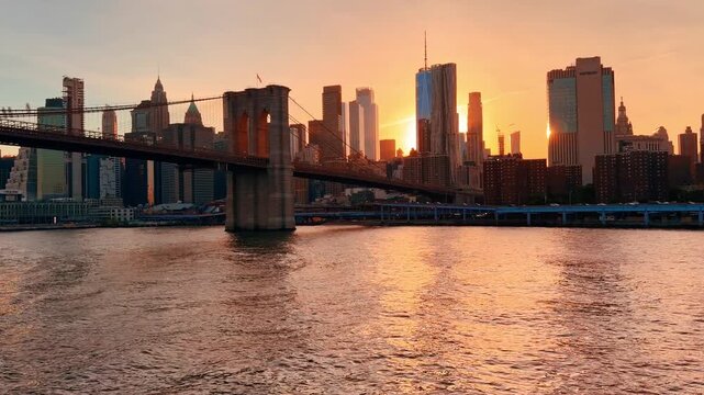 New York, USA, 22 May 2026: Brooklyn Bridge and Manhattan skyline at golden sunset. Warm orange light behind towers with sun reflections across East River surface.