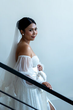 Beautiful Asian bride in elegant white wedding dress with veil posing on staircase, looking at camera.