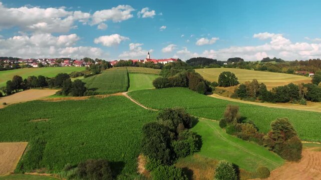 Flying towards a village on the horizon on beautiful green hills in Bavaria, Germany, with trees, blue sky and fluffy white clouds in the background