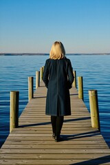 Fototapeta premium Blond woman stands on wooden pier looking at vast blue water. She wears long denim coat and black boots. Calm seascape and clear sky. Enjoying nature and quiet time.