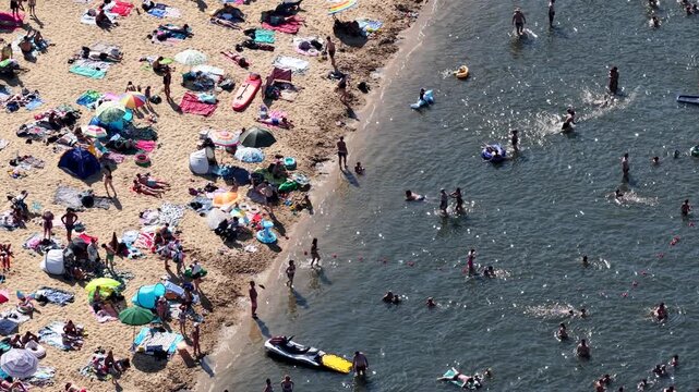 Aerial drone view of a crowded summer lake beach with people swimming and sunbathing. Colorful umbrellas, water obstacle course on lake, on a sunny day. Lake Pogoria in Dabrowa Gornicza, Poland
