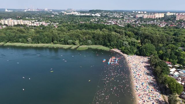 Aerial drone view of a crowded summer lake beach with people swimming and sunbathing. Colorful umbrellas, water obstacle course on lake, on a sunny day. Lake Pogoria in Dabrowa Gornicza, Poland