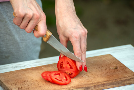 Person's hands holding a knife, slicing a ripe red tomato into thin pieces on a wooden cutting board, preparing ingredients for a healthy meal