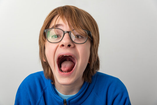 Boy with glasses and blue jacket yelling toward the camera, mouth wide open in a vivid expression of surprise, fear or excitement against a clean white background