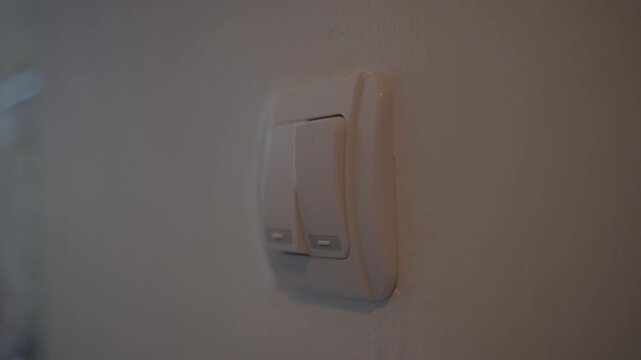 Close-up of a hand pressing a light switch on a white wall, turning on the room light as part of an everyday home routine. Turn on room electric light wall switch pushing button.