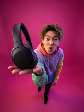 Young Asian man holding out black headphones on pink backdrop