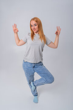 Woman standing on one leg, balancing while smiling at the camera and holding up three fingers on one hand and making an OK gesture with the other hand
