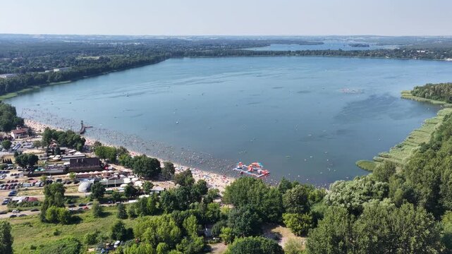 Aerial drone view of a crowded summer lake beach with people swimming and sunbathing. Colorful umbrellas, water obstacle course on lake, on a sunny day. Lake Pogoria in Dabrowa Gornicza, Poland