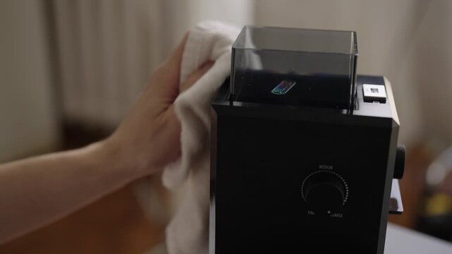 Close-up view of person hands using white cloth to wipe down modern black electric burr coffee grinder, performing regular maintenance for optimal performance and cleanliness. Shooting in slow motion
