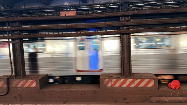 Moving subway train with motion blur, New York City. Silver car streaking past steel columns with striped barriers and industrial ceiling.