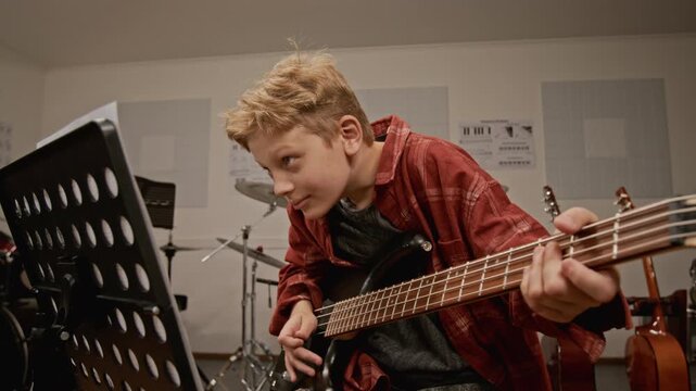 Caucasian teenage boy practicing electric bass guitar in music classroom, looking at music stand. Footage for promoting music schools, youth education, and creative hobbies