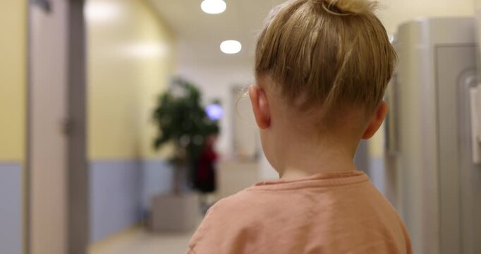 Cute little girl with blonde hair tied in a bun drinking water from a paper cup to quench her thirst in a public building hallway, then she turns and walks away from the camera